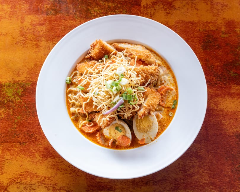 A white bowl filled with a rich orange curry, noodles, fried chicken, sliced eggs, and vegetables.