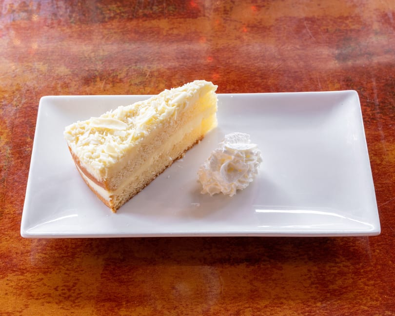 Slice of white chocolate cake with whipped cream on a white rectangular plate.