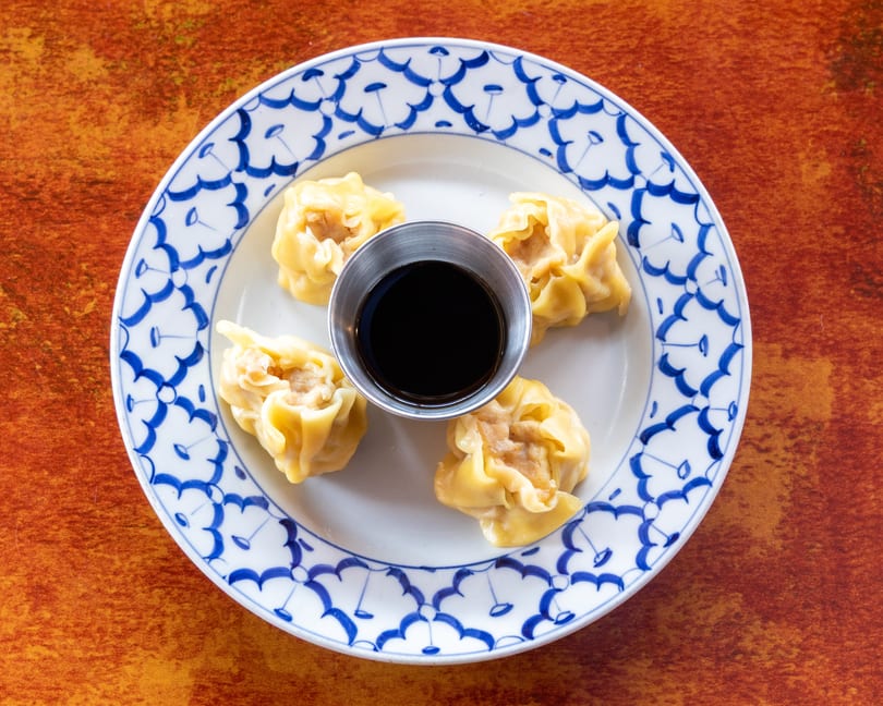 Four steamed dumplings with a small cup of soy sauce on a white and blue patterned plate.